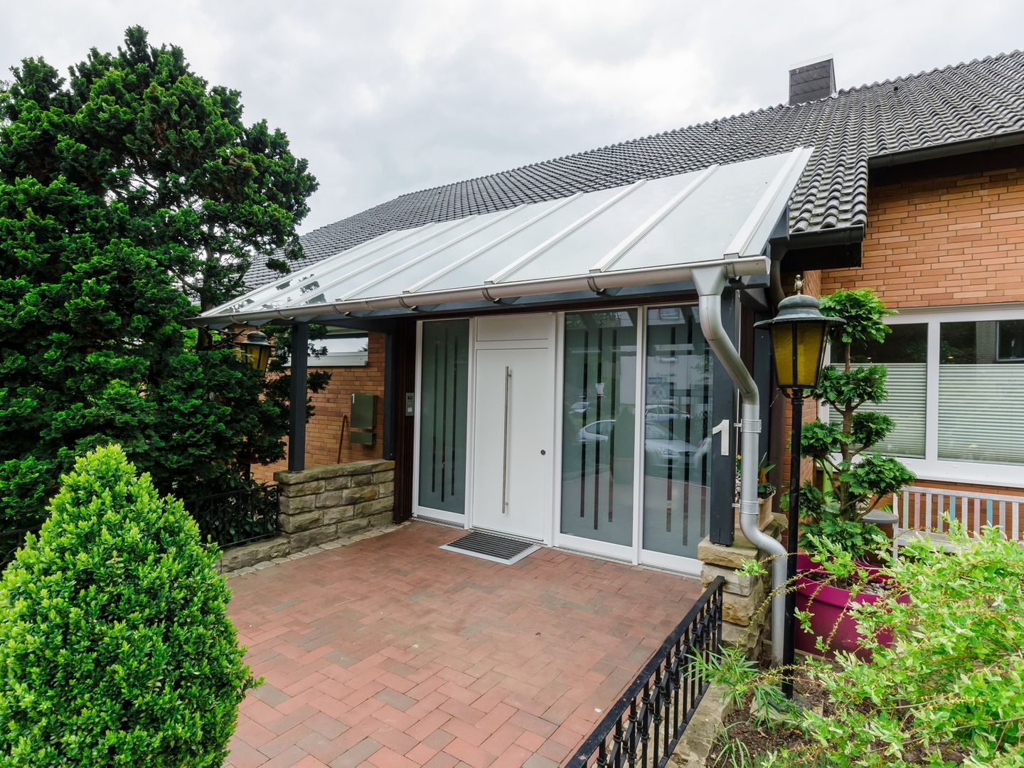 Hauseingang mit Glasvordach, weißer Tür und Grünpflanzen